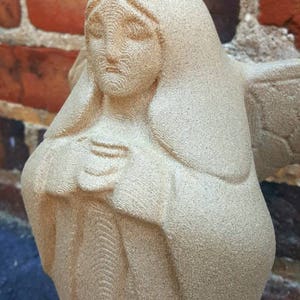 May include: A beige stone sculpture of a figure with a serene expression, wearing a draped garment and a head covering. The sculpture is set against a backdrop of a red brick wall.