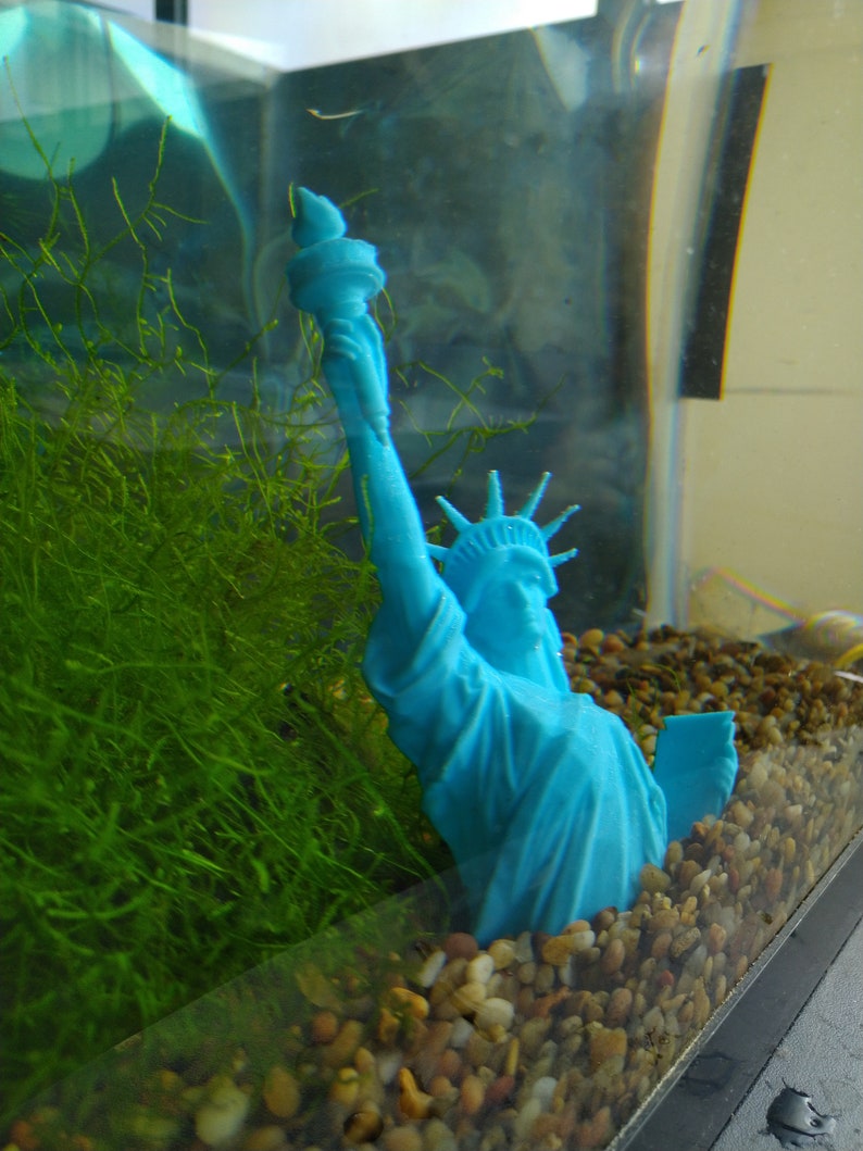 K&ouml;nnte beinhalten: Eine blaue Plastikstatue der Freiheitsstatue in einem Aquarium mit gr&uuml;nen Pflanzen und Kies.