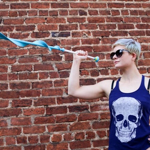 Puede incluir: Una mujer con una camiseta azul de tirantes con un estampado de calavera blanca y gafas de sol sostiene un juguete azul y blanco. El fondo es una pared de ladrillo rojo.