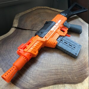 May include: An orange and gray toy blaster gun with the text "AEON" on the side. The toy gun has a gray magazine, a black handle, and an orange barrel with holes. The toy is made of plastic.