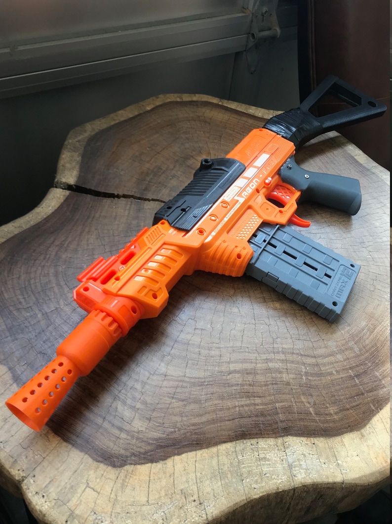 May include: An orange and gray toy blaster gun with the text "AEON" on the side. The toy gun has a gray magazine, a black handle, and an orange barrel with holes. The toy is made of plastic.