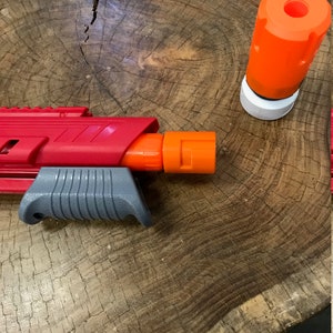 May include: A red and gray toy gun with an orange barrel and a separate orange barrel attachment. The gun is lying on a wooden surface.