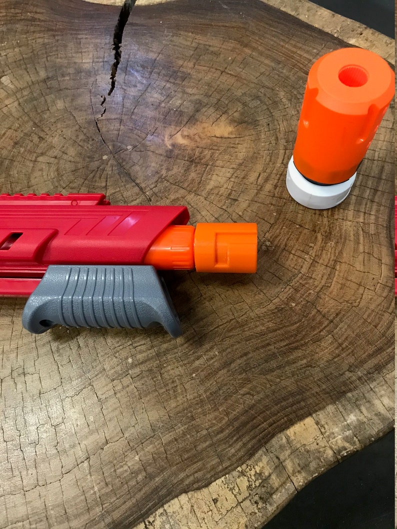 May include: A red and gray toy gun with an orange barrel and a separate orange barrel attachment. The gun is lying on a wooden surface.