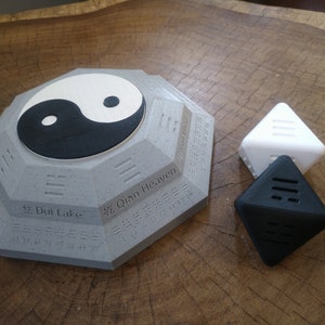 May include: A gray octagonal base with a yin yang symbol on top. The base has Chinese characters and numbers around the edge. There are two triangular pieces, one white and one black, with lines on them.
