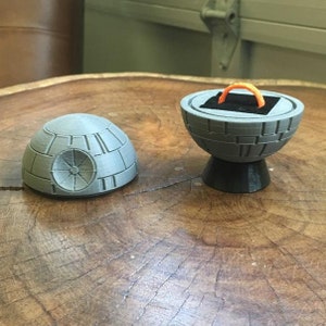 May include: A gray and black 3D printed ring box shaped like the Death Star from Star Wars. The box is open and a gold ring is inside.
