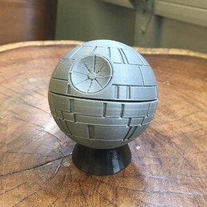 May include: A gray 3D printed Death Star model with a black base. The model is a replica of the iconic spaceship from the Star Wars franchise.