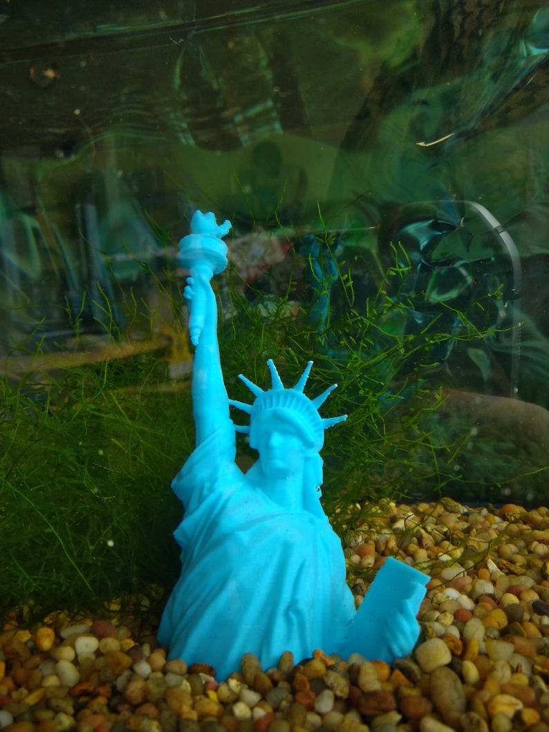 K&ouml;nnte beinhalten: Eine blaue Plastikstatue der Freiheitsstatue, die eine Fackel h&auml;lt, ist teilweise in einem mit Kies und gr&uuml;nen Wasserpflanzen gef&uuml;llten Aquarium versunken.