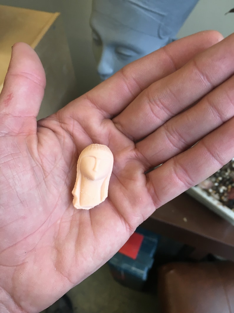 May include: A small, pink, ceramic figurine of a woman's head with long hair.