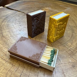 May include: Two matchboxes with a book-like design. One is brown with an embossed design and the other is gold with the text "Spell Book". The brown matchbox is open and shows a row of matches with green tips.