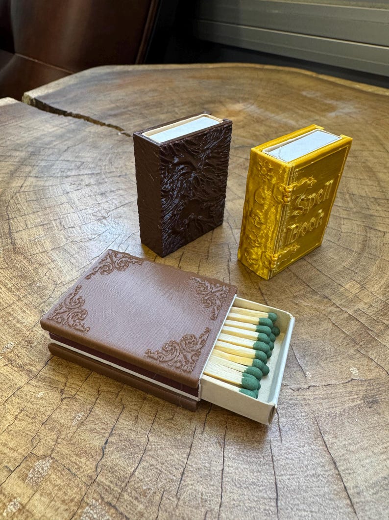 May include: Two matchboxes with a book-like design. One is brown with an embossed design and the other is gold with the text "Spell Book". The brown matchbox is open and shows a row of matches with green tips.