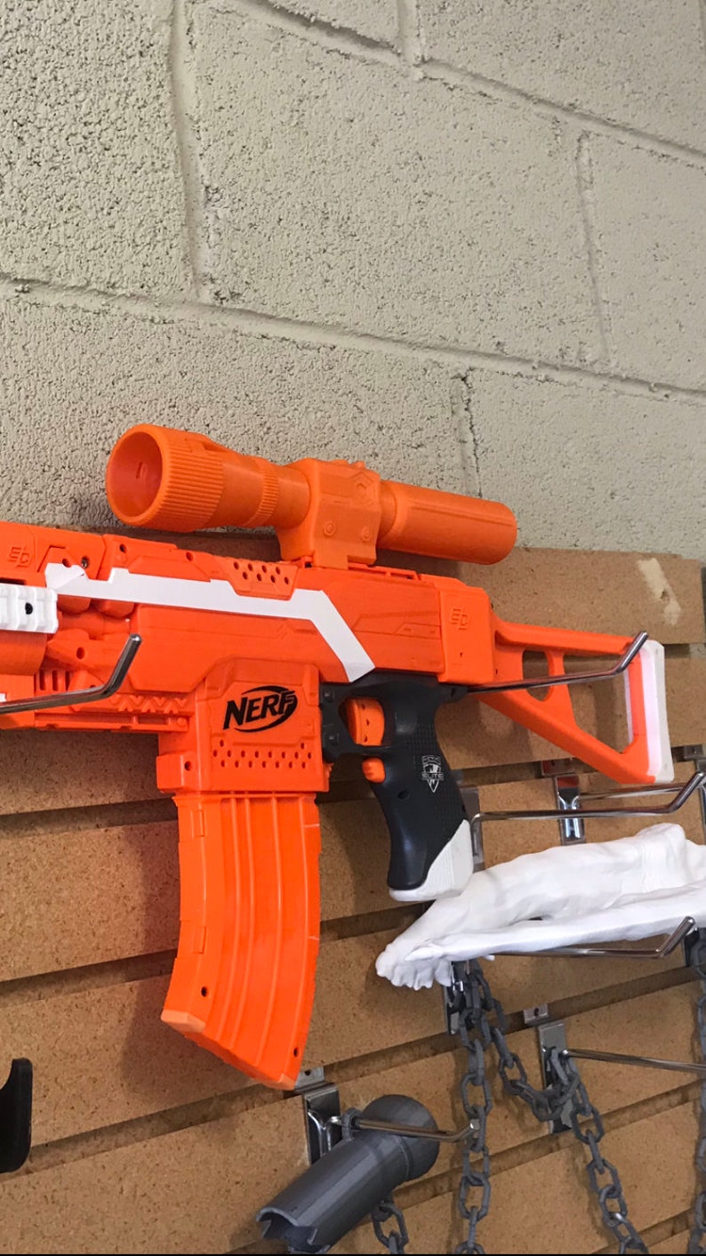 May include: An orange and white Nerf gun with a black scope and a large orange magazine. The gun is mounted on a wall with a chain and hook.