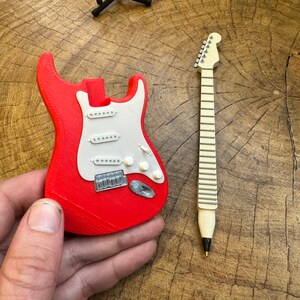 May include: A red and white miniature electric guitar with a black guitar stand in the background. The guitar is made of plastic and has a detailed design.