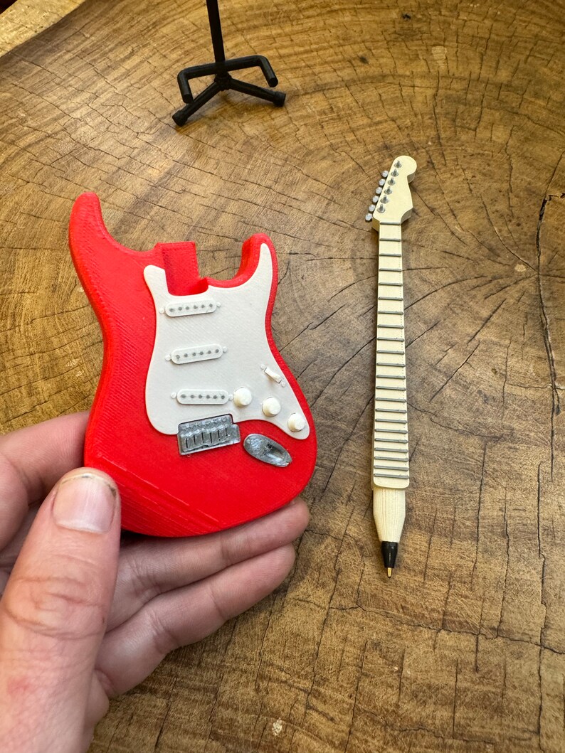 May include: A red and white miniature electric guitar with a black guitar stand in the background. The guitar is made of plastic and has a detailed design.