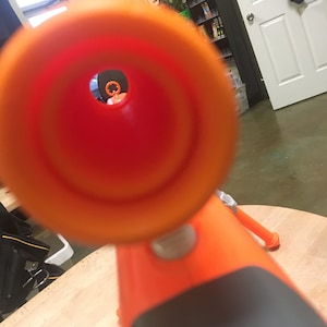 May include: Close-up view through the orange barrel of a toy gun. The barrel is pointed at a blurry background.