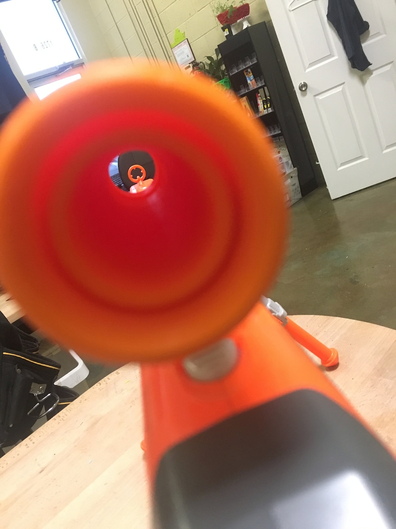 May include: Close-up view through the orange barrel of a toy gun. The barrel is pointed at a blurry background.