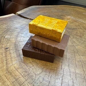 May include: Three decorative books stacked on top of each other. The top book is gold with the title "Spell Book" embossed on the cover. The middle book is brown with a decorative design. The bottom book is dark brown with a textured surface.