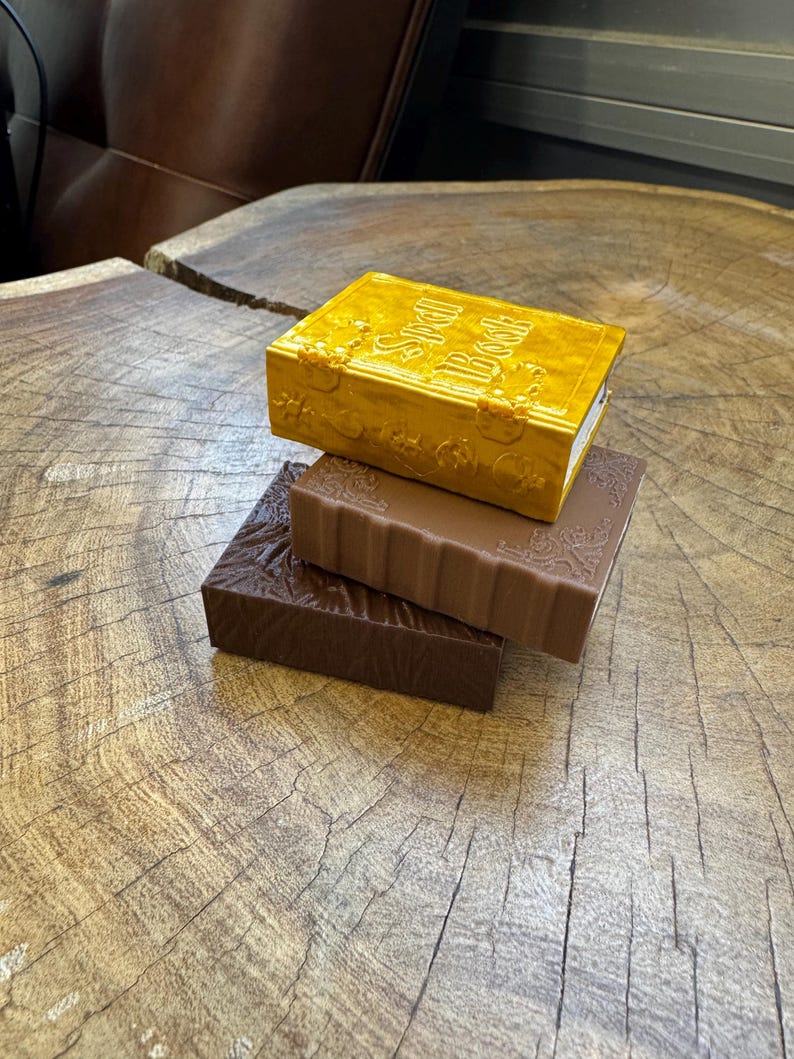 May include: Three decorative books stacked on top of each other. The top book is gold with the title "Spell Book" embossed on the cover. The middle book is brown with a decorative design. The bottom book is dark brown with a textured surface.