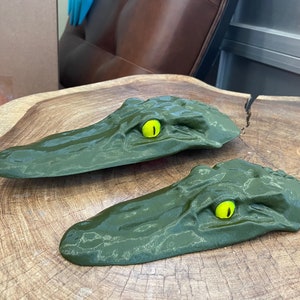 May include: Two green alligator head shaped wall mounts with yellow eyes. The alligator heads are made of a textured material and are mounted on a wooden surface.