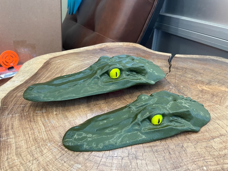 May include: Two green alligator head shaped wall mounts with yellow eyes. The alligator heads are made of a textured material and are mounted on a wooden surface.