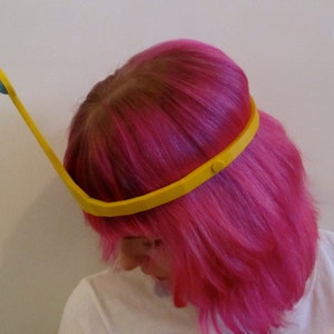 May include: A yellow headband with a blue orb on the end. The headband is worn by a person with pink hair.