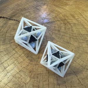 May include: Two geometric octahedron structures made of translucent white material. Each frame contains black triangular components and smooth, light blue and gray stones held within the open lattice framework of the polyhedral shapes.