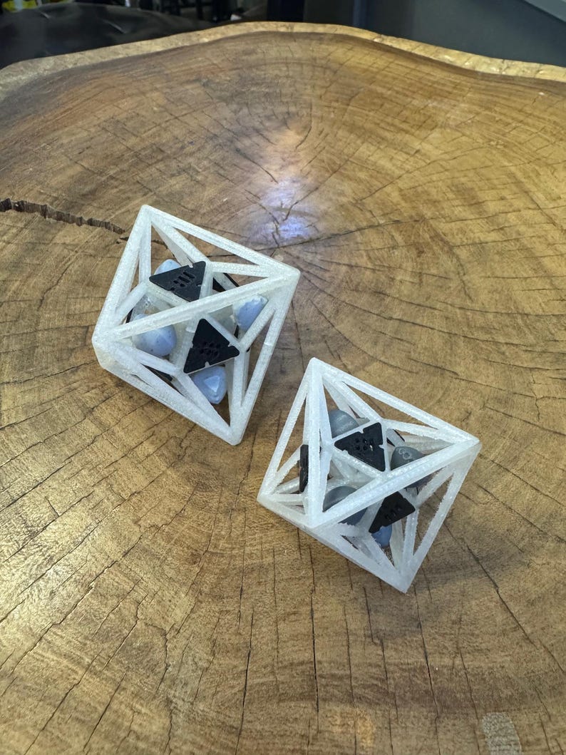 May include: Two geometric octahedron structures made of translucent white material. Each frame contains black triangular components and smooth, light blue and gray stones held within the open lattice framework of the polyhedral shapes.