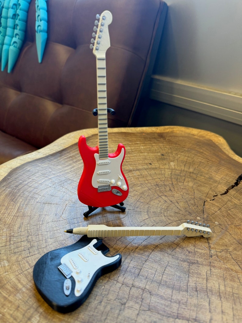 May include: Two miniature electric guitars, one red with a white pickguard and one black with a white pickguard. The guitars are on a wooden surface.