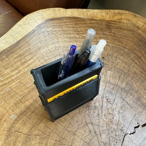 May include: A black and yellow 3D printed pen holder with four pens inside. The pens are blue, black, and clear.