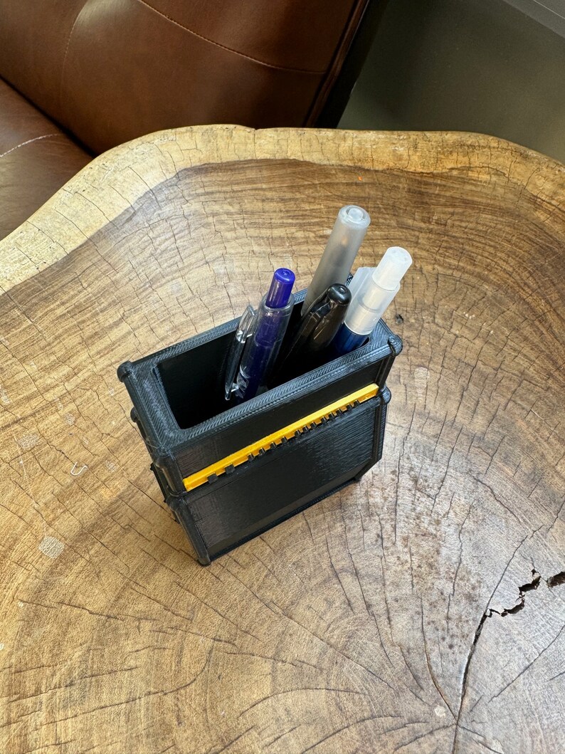 May include: A black and yellow 3D printed pen holder with four pens inside. The pens are blue, black, and clear.