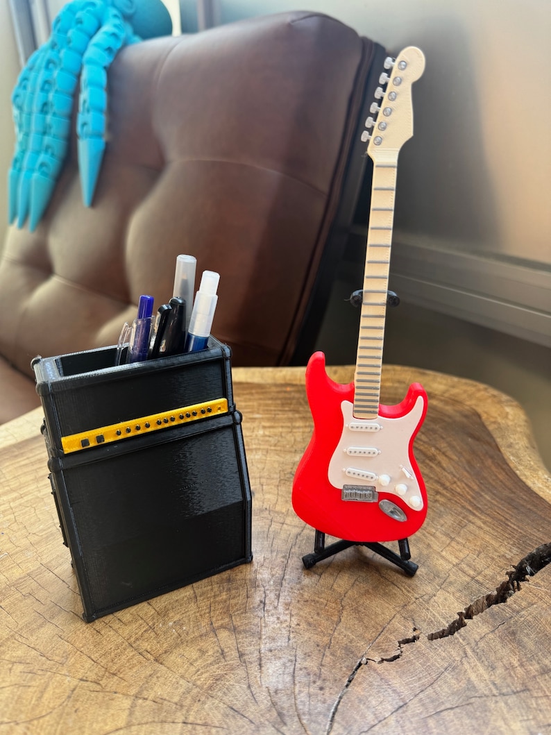 May include: A black and yellow guitar amplifier-shaped pencil holder with pens and markers inside, next to a red electric guitar on a stand. The guitar has a white pickguard and a wooden neck.