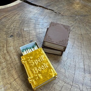 May include: A gold matchbox with the words "Spell Book" on the cover. The matchbox is sitting on a wooden surface next to a brown matchbox.