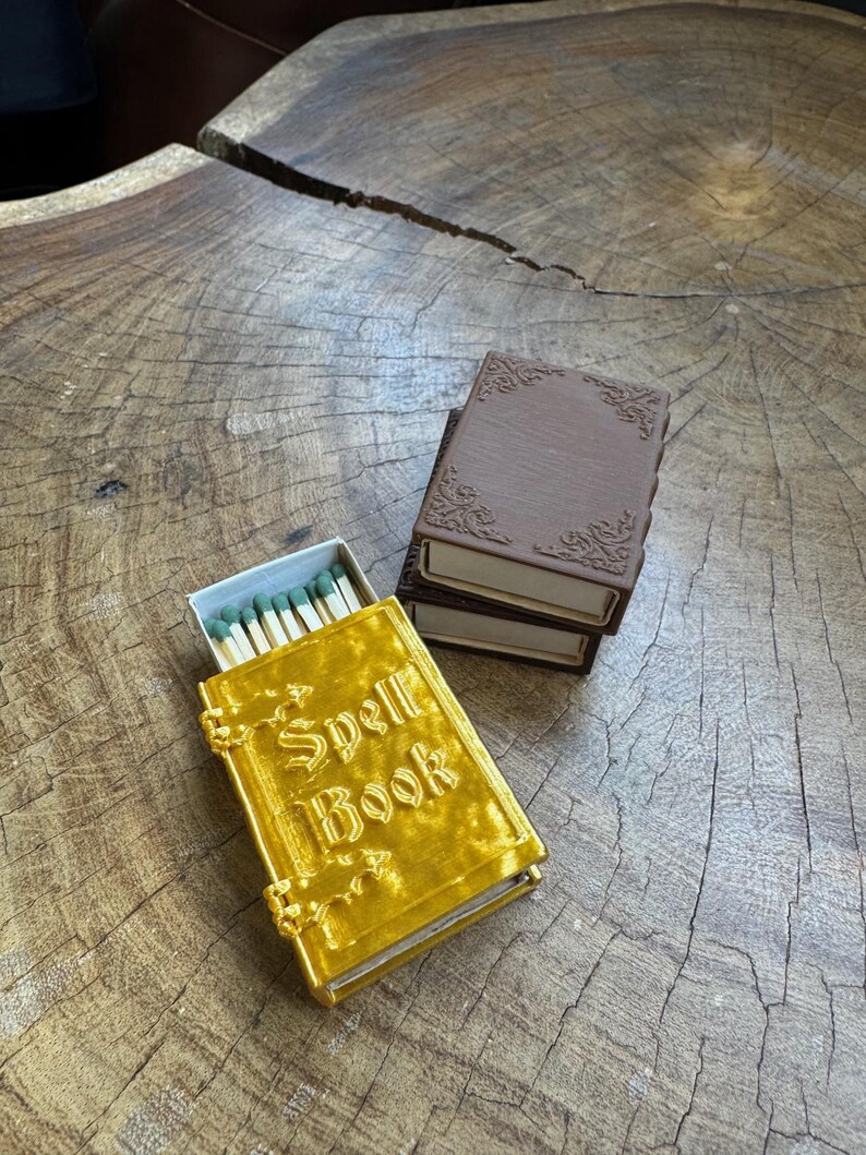 May include: A gold matchbox with the words "Spell Book" on the cover. The matchbox is sitting on a wooden surface next to a brown matchbox.