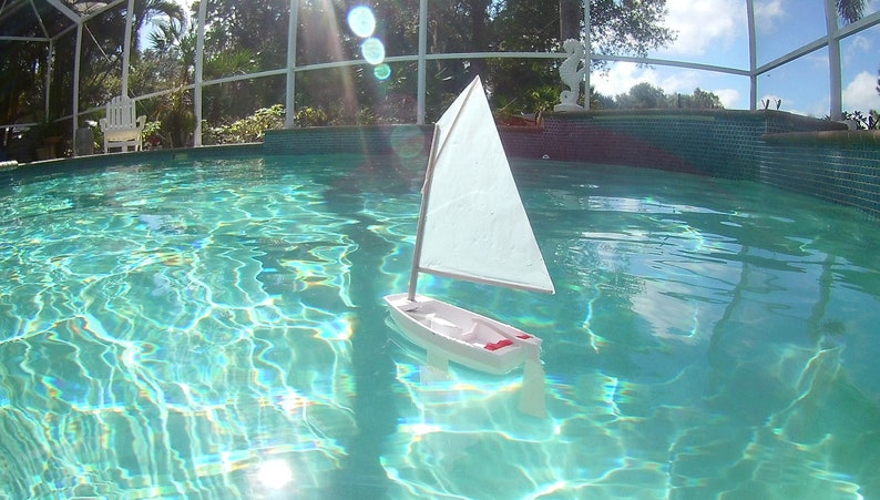 Puede incluir: Un velero de juguete blanco con una vela blanca flota en una piscina azul. La piscina est&aacute; rodeada por un patio cubierto.
