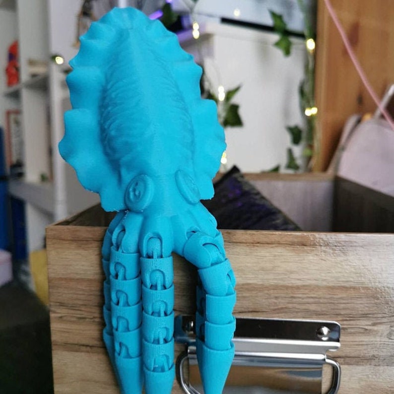 May include: A turquoise blue 3D printed octopus figurine with multiple tentacles. The octopus is perched on the edge of a wooden drawer with a silver handle.