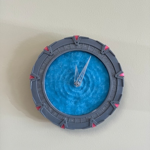 May include: A grey and red clock shaped like the Stargate from the TV show Stargate SG-1. The clock face is blue with a swirling pattern and a single silver hand.