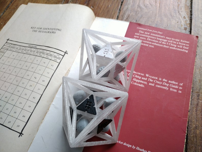 May include: Two white 3D-printed geometric cage structures, each containing several small gray spheres and one triangular piece. One cage holds a black triangular piece with six dots, while the other holds a white triangular piece with six dots.