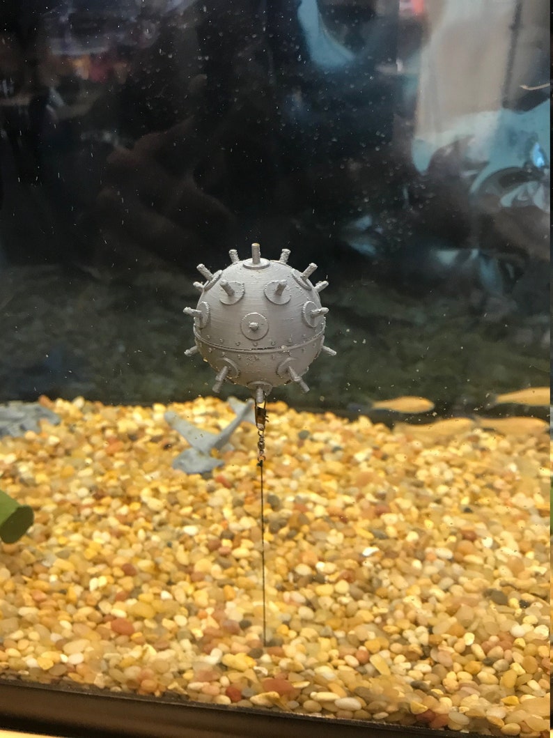 May include: A gray, spherical, metal object with a chain attached. The object has a textured surface and is covered in small, round protrusions. The object is suspended in a clear tank filled with small, round, yellow and brown stones.