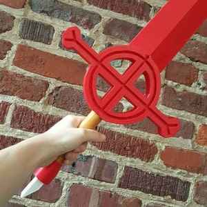 Op de afbeelding: Een rood plastic zwaard met een cirkelvormig ontwerp op het blad en een houten handvat. Het zwaard wordt in de hand van een persoon gehouden.