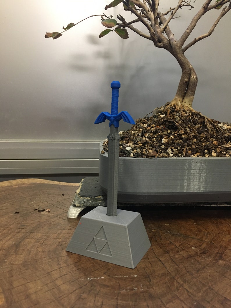 May include: A blue plastic sword with a gold handle and a blue and gold winged design. The sword is standing upright in a gray plastic stand with a triangular design.