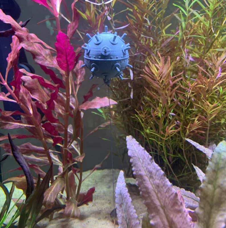 May include: A gray, 3D printed, spiky, round ornament hanging in a fish tank with green and red plants.