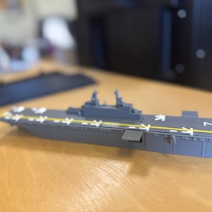 May include: A grey model aircraft carrier with white markings and yellow stripes. The model has a flat top with the number 7 on the side. It is on a wooden surface.