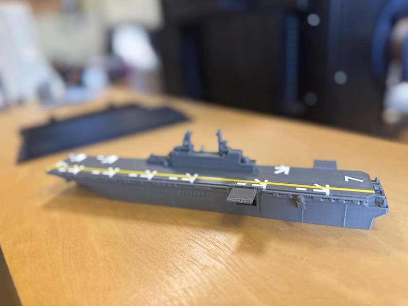 May include: A grey model aircraft carrier with white markings and yellow stripes. The model has a flat top with the number 7 on the side. It is on a wooden surface.