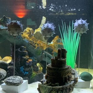 May include: Three gray, spiky, round ornaments hang from chains in a fish tank. The ornaments are suspended above a dark gray, detailed model of a tugboat. The tank also contains green plastic plants and a gray rock.