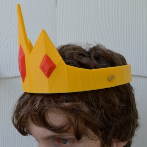 May include: A yellow plastic crown with red diamond-shaped accents. The crown is worn on a person's head.