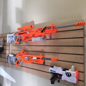 May include: Three toy blasters mounted on a wall. The top blaster is orange and gray with the text "RAPTORSTRIKE N-STRIKE" on the side. The middle blaster is orange and gray with the text "NERF  FLASHFIRE" on the side. The bottom blaster is white and black with the text "RIVAL" on the side.