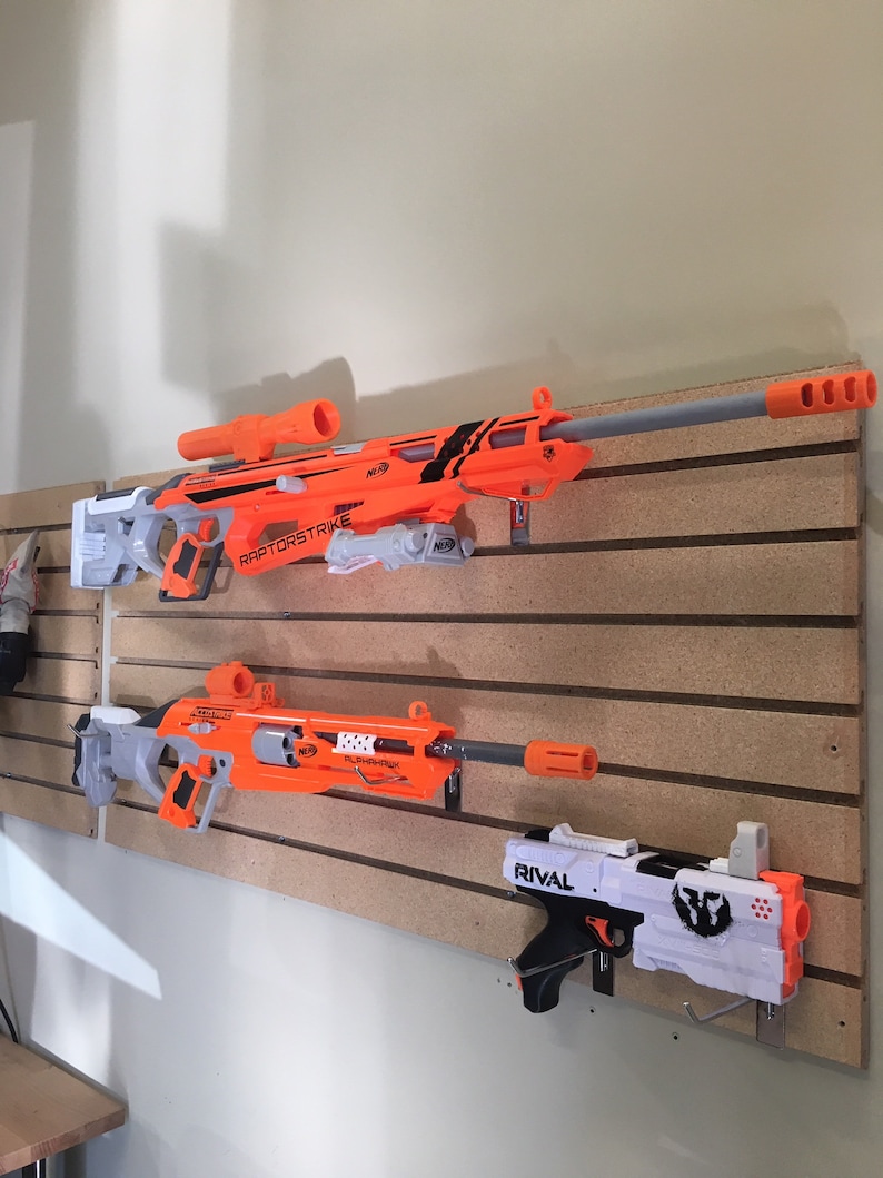 May include: Three toy blasters mounted on a wall. The top blaster is orange and gray with the text "RAPTORSTRIKE N-STRIKE" on the side. The middle blaster is orange and gray with the text "NERF  FLASHFIRE" on the side. The bottom blaster is white and black with the text "RIVAL" on the side.