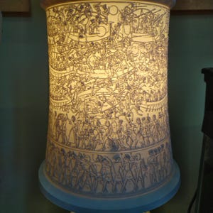 Pu&ograve; includere: Un paralume in ceramica bianca con un design intricato di persone e animali. Il paralume &egrave; illuminato dall'interno, proiettando una calda luce.