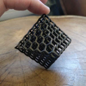 Puede incluir: Un objeto geom&eacute;trico de color negro impreso en 3D con un patr&oacute;n de panal. El objeto est&aacute; hecho de pl&aacute;stico y tiene una base cuadrada.