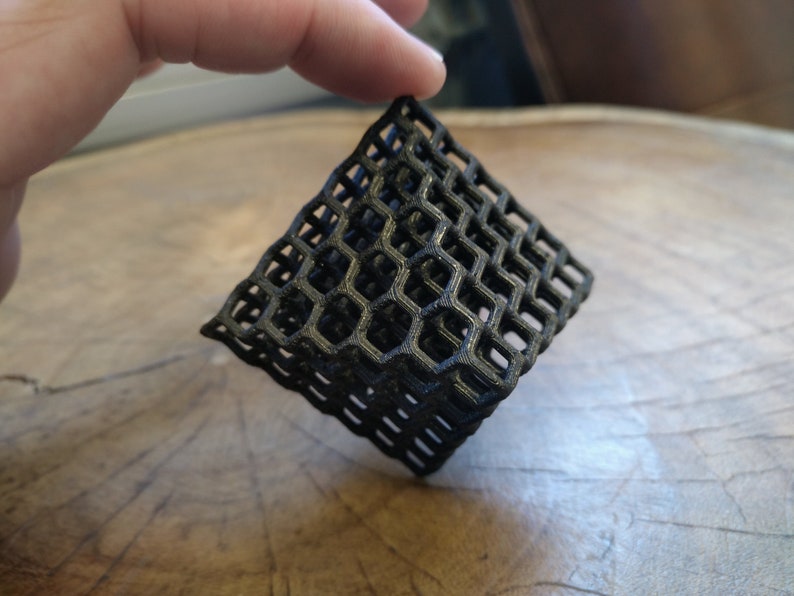 Puede incluir: Un objeto geom&eacute;trico de color negro impreso en 3D con un patr&oacute;n de panal. El objeto est&aacute; hecho de pl&aacute;stico y tiene una base cuadrada.