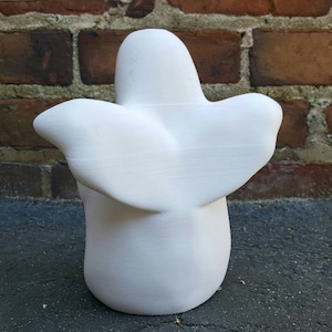 K&ouml;nnte beinhalten: Wei&szlig;e Keramik-Geisterfigur mit einer glatten, abgerundeten Form und einem fl&uuml;gelartigen Design auf der R&uuml;ckseite. Der Geist steht auf einer dunklen Oberfl&auml;che.